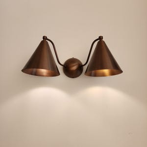 May include: A bronze-colored wall sconce with two conical shades. The shades are angled downward, and the fixture is mounted on a round base. The sconce is made of metal and has a warm, inviting glow.