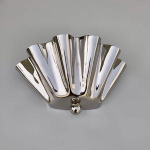May include: A polished silver-colored decorative wall sconce with a fan-shaped design. The sconce has a series of vertical, curved ridges and a small, spherical accent at the base. The reflective surface creates a play of light and shadow.