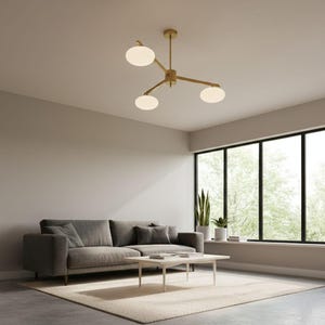 Puede incluir: Una lámpara de araña moderna en tono dorado con tres pantallas esféricas de vidrio esmerilado. La lámpara está suspendida del techo, sobre un salón con un sofá gris, una alfombra y una mesa de centro.