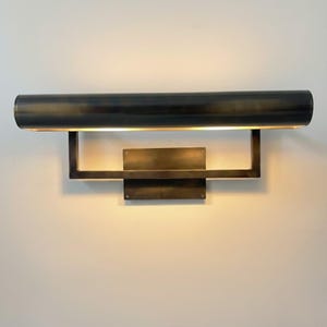 Puede incluir: Aplique de pared de bronce oscuro con una placa trasera rectangular y una luminaria cilíndrica horizontal. La luminaria está soportada por un marco rectangular. El aplique emite un brillo cálido y acogedor.