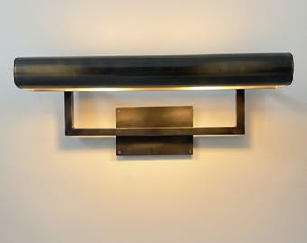 May include: A dark bronze wall sconce with a rectangular backplate and a horizontal, cylindrical light fixture. The light fixture is supported by a rectangular frame. The sconce emits a warm, inviting glow.