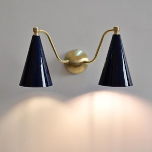 May include: A wall light with two navy blue, cone-shaped shades. The fixture has a gold-coloured central mount and curved arms. The light shines downwards, illuminating the wall below.