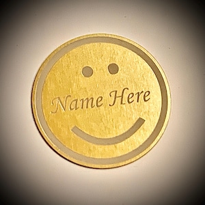 Op de afbeelding: Een ronde, goudkleurige smiley met de tekst "Name Here" in het midden gegraveerd. Het ontwerp heeft twee kleine stippen voor ogen en een gebogen lijn voor de mond. De buitenrand heeft een lichtere rand.