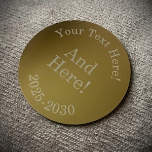 May include: A gold-colored, circular item with engraved text. The text reads "Your Text Here!" around the top and "And Here!" in the center. The numbers "2025-2030" are at the bottom. The item is on a gray, textured surface.