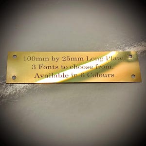 May include: A rectangular brass plate with rounded corners and four holes. The plate measures 10cm by 2.5cm and features engraved text: "100mm by 25mm Long Plate, 3 Fonts to choose from, Available in 6 Colours."