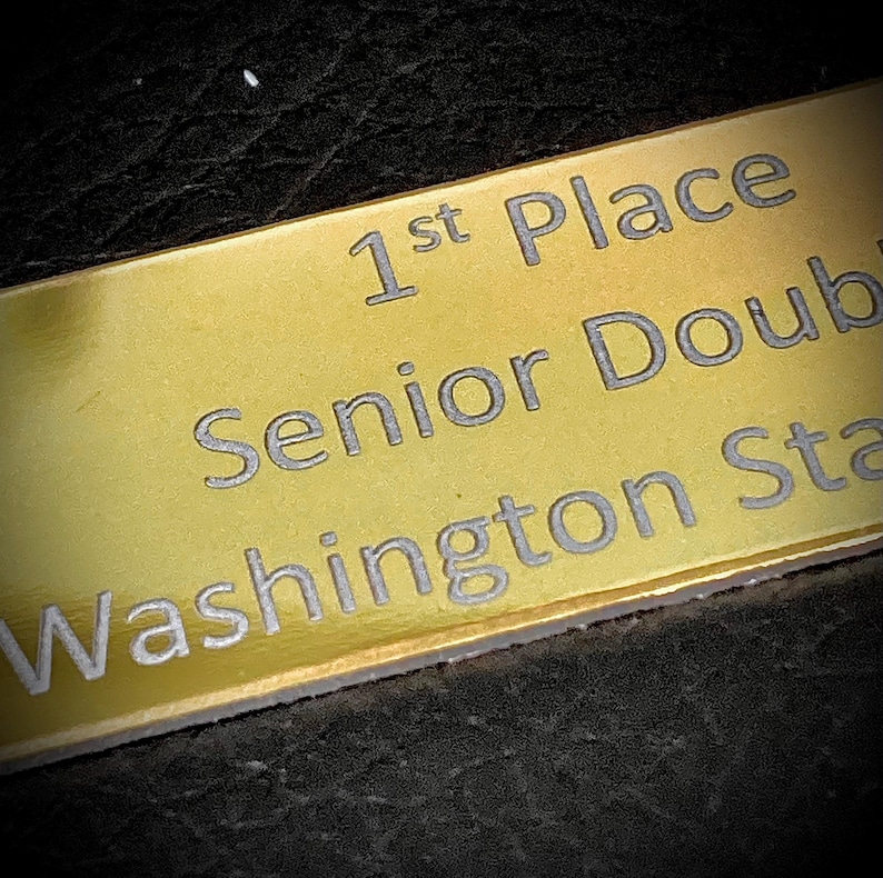 K&ouml;nnte beinhalten: Goldfarbene Plakette mit silberner Schrift: "1st Place Senior Doub Washington Sta". Die rechteckige Plakette ist vermutlich aus Metall. Der dunkle, strukturierte Hintergrund hebt die Plakette hervor.