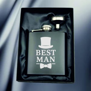 May include: A black flask with a silver cap and a small funnel, presented in a black box. The flask has the words "BEST MAN" and a top hat and bow tie design in white.