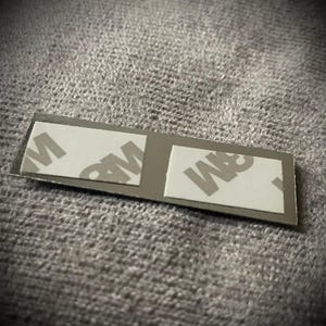 K&ouml;nnte beinhalten: Ein kleiner, rechteckiger, silberfarbener Klebestreifen mit zwei wei&szlig;en Klebepads. Auf den Pads ist das Logo "3M" aufgedruckt. Der Streifen ist f&uuml;r Klebe- oder Montageanwendungen konzipiert.