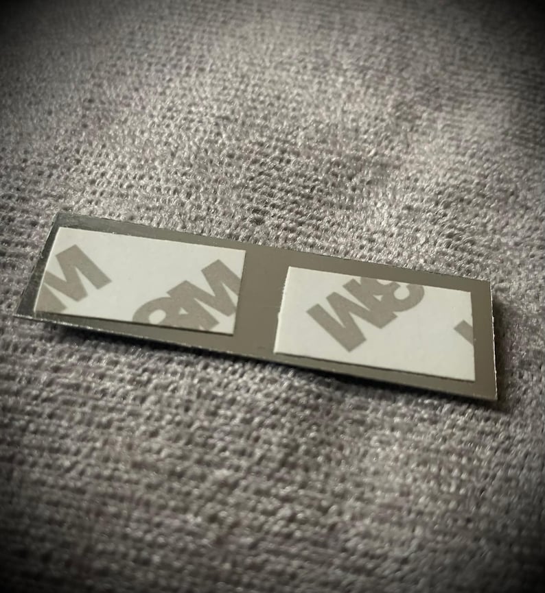 K&ouml;nnte beinhalten: Ein kleiner, rechteckiger, silberfarbener Klebestreifen mit zwei wei&szlig;en Klebepads. Auf den Pads ist das Logo "3M" aufgedruckt. Der Streifen ist f&uuml;r Klebe- oder Montageanwendungen konzipiert.