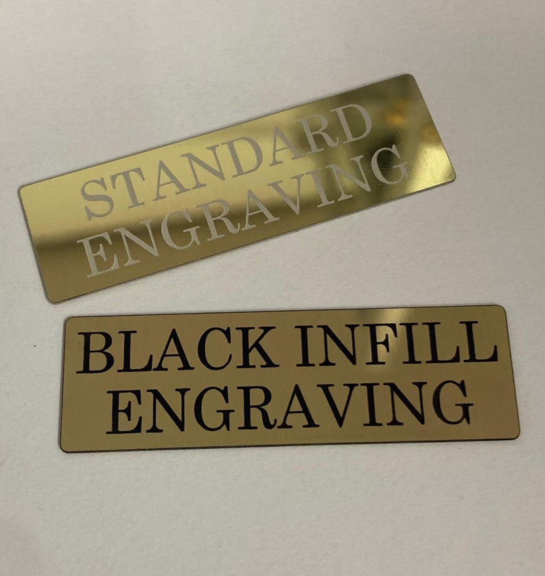 K&ouml;nnte beinhalten: Zwei rechteckige, goldfarbene Metallplatten mit graviertem Text. Die obere Platte tr&auml;gt die Aufschrift "STANDARD ENGRAVING" in heller Farbe. Die untere Platte tr&auml;gt die Aufschrift "BLACK INFILL ENGRAVING" in Schwarz.
