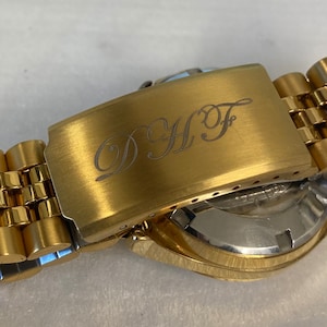 Pu&ograve; includere: Un orologio dorato con un cinturino in metallo dorato. Il cinturino ha una fibbia con le iniziali "DHF" incise.