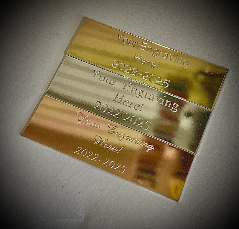 K&ouml;nnte beinhalten: Drei rechteckige Metallplatten in Gold-, Silber- und Kupfert&ouml;nen. Jede Platte tr&auml;gt die Gravur "Your Engraving Here!" und den Zeitraum "2022-2025". Die Platten sind leicht &uuml;berlappend angeordnet.