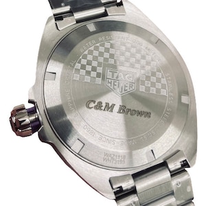 Pu&ograve; includere: Un orologio Tag Heuer argentato con un cinturino in acciaio inossidabile. Il fondello dell'orologio presenta un motivo a scacchi e la scritta "C&M Brown" incisa.