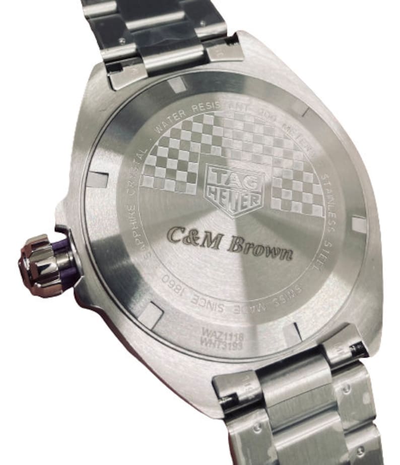 Pu&ograve; includere: Un orologio Tag Heuer argentato con un cinturino in acciaio inossidabile. Il fondello dell'orologio presenta un motivo a scacchi e la scritta "C&M Brown" incisa.