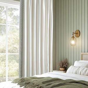 May include: A bedroom with a white bed with a green throw blanket, a woven headboard, and a woven nightstand. The walls are painted a light green and there are two brass wall sconces with clear glass globes. A framed print of a beach scene hangs on the wall.