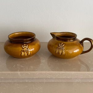 May include: A ceramic sugar bowl and creamer set in a glossy mustard-brown glaze. Both pieces feature a raised emblem with the text 1650 and a crown motif. The creamer has a small pouring spout and a curved handle, while the sugar bowl is open.