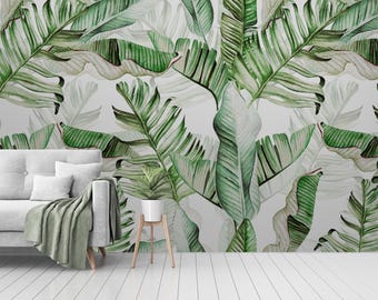 Op de afbeelding: Een witte muur met een groene en witte behang met tropische bladmotieven. Voor de muur staat een witte bank met een groene deken. Een kleine groene plant staat op een houten standaard.