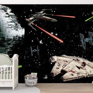Könnte beinhalten: Ein Wandbild mit einer Weltraumschlacht-Szene, das den Todesstern, einen X-Wing-Sternenjäger mit roten Lasern, TIE-Jäger mit grünen Lasern und den Millennium Falken vor einem schwarzen Sternenhimmel zeigt, platziert hinter einem weißen Kinderbett.