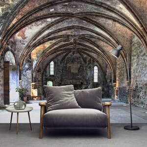 May include: Interior scene with a grey velvet armchair, wooden armrests, a small round side table, and a black floor lamp. The backdrop is a historic stone building with arched ceilings and windows.