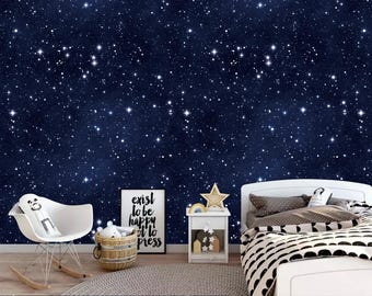 May include: A white rocking chair with a white stuffed animal in the seat. A white nightstand with a gold star on top sits next to a white bed with black and white bedding. A framed print with the text "exist to be happy or to press" is on the wall behind the chair. The wall is covered in a dark blue wallpaper with white stars.