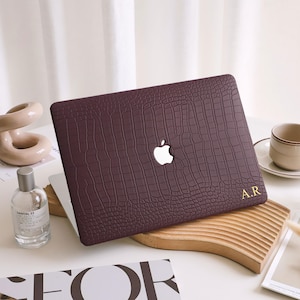 May include: A burgundy laptop case with a crocodile skin texture. The case has a white Apple logo and gold initials "A.R." on the lower right corner. The case is on a wooden cutting board.
