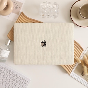 May include: A cream-colored laptop with a textured knit pattern cover, featuring the Apple logo. The laptop is placed on a wooden tray, surrounded by a keyboard, perfume bottle, and decorative items. The overall aesthetic is minimalist and neutral.