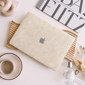 Könnte beinhalten: Cremefarbene Laptop-Hülle mit strukturiertem Muster und Apple-Logo. Die Initialen "B.C" sind goldfarben aufgedruckt. Die Hülle liegt auf einem Holztablett, mit einem Buch und einer Parfümflasche daneben. Der Buchtitel lautet "DA FITZ".