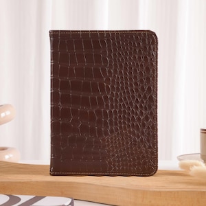 Puede incluir: Una funda rectangular marrón para tableta con textura de piel de cocodrilo. La funda está hecha de un material marrón oscuro y tiene un borde cosido. La funda está de pie sobre una superficie de madera.