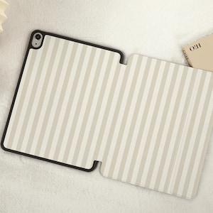 Può includere: Una custodia per iPad a righe beige e bianche con un bordo nero. La custodia è aperta e ripiegata all'indietro, mostrando il rivestimento interno.