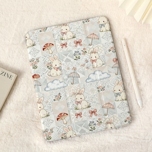 May include: A light-colored tablet case featuring a repeating pattern of bunnies, mushrooms, umbrellas, and clouds. The case has a patchwork design with floral accents and pink bows. A white stylus and a magazine are also visible.