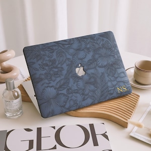May include: A blue laptop case with a floral design and the initials "NS." embossed in gold. The case has a textured surface and a subtle sheen.