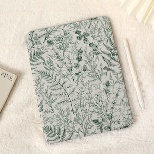 Peut inclure: Une coque d'iPad blanche avec un imprimé botanique vert représentant des fougères et d'autres feuillages. La coque a une surface texturée subtile.