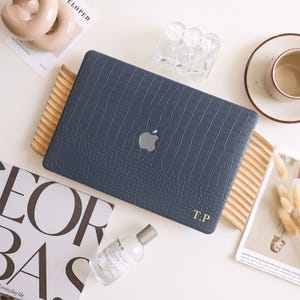 May include: A navy blue laptop case with a crocodile skin texture, featuring a silver Apple logo. The case rests on a wooden tray, with the initials "T.P" in gold. Other items include a perfume bottle and a decorative object.