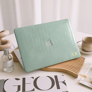 Puede incluir: Una funda para portátil verde menta con textura de piel de cocodrilo. La funda tiene un recorte para el logotipo de Apple y las iniciales doradas "A.R." en la esquina. La funda está sobre una mesa blanca con otros objetos decorativos.