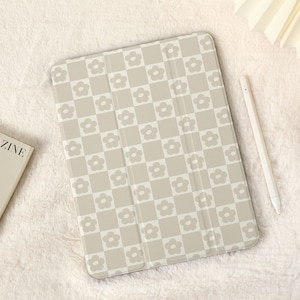 May include: A beige iPad case with a white checkered pattern and small white flowers. The case is closed and lying on a white fluffy surface.