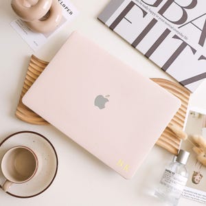 Puede incluir: Un portátil rosa claro con el logotipo de Apple, apoyado en una bandeja de madera. Las iniciales "B.K" están impresas en amarillo en el portátil. También se ven una taza y un platillo, un frasco de perfume y una revista.