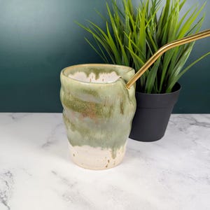 May include: A handcrafted ceramic mug with a distinctive, textured pattern. The mug has a light green glaze that blends into a creamy white at the bottom. A gold-coloured metal straw extends from the side. A small black pot with green plants is in the background.