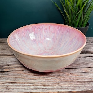 Puede incluir: Un cuenco de cerámica con interior rosa y blanco y exterior beige claro. El cuenco tiene una forma ancha y poco profunda y se asienta sobre una superficie de madera. El interior tiene un efecto veteado.