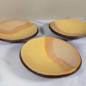 Op de afbeelding: Drie kleine, ronde keramische schalen met een bruine rand en een gele en beige glazuur. De schalen zijn in een verspringend patroon op een wit oppervlak gerangschikt.