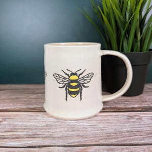 May include: Cream ceramic mug with a black and yellow bee design. The mug has a curved handle and a flared base. The bee illustration is centered on the front. The mug is displayed on a wooden surface.