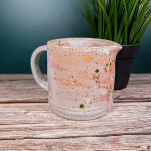 Op de afbeelding: Handgemaakte keramische kan met een handvat en tuit. De kan heeft een gespikkelde roze en crèmekleurige glazuur met oranje en groene accenten. De kan staat op een houten oppervlak.