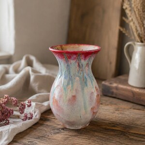 May include: A ceramic vase with a flared rim, featuring a reactive glaze finish that blends shades of vibrant red at the top, transitioning into streaks of light blue and soft cream, with a rounded body and a smooth, glossy surface texture.