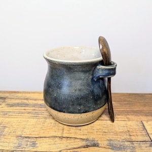 Peut inclure: Une tasse en céramique bleue et blanche avec une cuillère en bois reposant dans la poignée. La tasse a un glaçage moucheté et une forme arrondie.