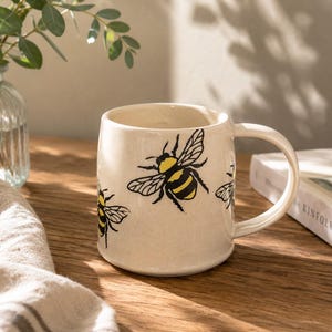 Può includere: Tazza in ceramica color crema con illustrazioni di api nere e gialle. La tazza ha una forma arrotondata e un manico grande. Il disegno dell'ape è ripetuto intorno alla tazza. La tazza è su una superficie di legno.