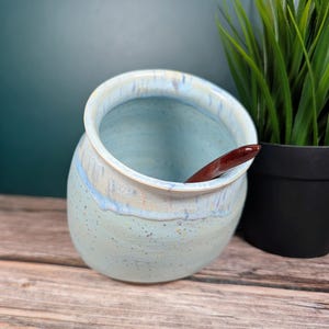 Può includere: Un vaso di ceramica con interno ed esterno azzurro chiaro, macchiato di blu più scuro e giallo. Il bordo ha una glassa bianca e azzurra. Un cucchiaio di legno marrone scuro è all'interno del vaso. Una pianta verde in vaso è sullo sfondo.