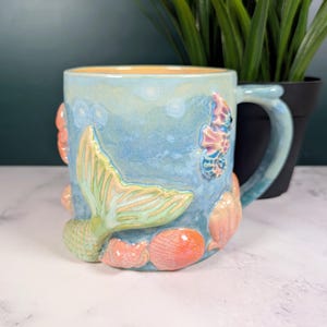 May include: Ceramic mug with a mermaid theme. The mug is light blue with a yellow interior and features a mermaid tail, seahorse, and seashells in shades of orange, pink, and green. The handle is light blue.