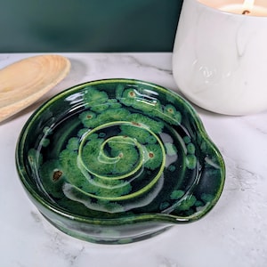 Może przedstawiać: Ręcznie wykonana, ciemnozielona ceramiczna podstawka pod łyżkę z unikalnym spiralnym wzorem. Podstawka pod łyżkę ma błyszczące wykończenie i małą krawędź do trzymania przyborów. Ceramiczny element jest zaakcentowany jaśniejszymi zielonymi i brązowymi detalami.
