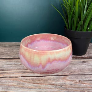 Op de afbeelding: Een handgemaakte keramische kom met een roze en gele glazuur. De kom heeft een afgeronde vorm en een gladde, glanzende afwerking. De kleuren vloeien samen in een aquareleffect. De kom wordt waarschijnlijk gebruikt voor het serveren van voedsel of als decoratief item.