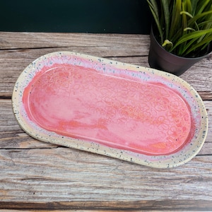 Puede incluir: Una fuente de cerámica hecha a mano con un diseño floral rosa. El plato ovalado tiene un borde beige moteado y un interior rosa brillante. La fuente es adecuada para servir aperitivos o postres.