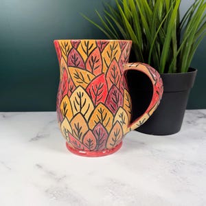 May include: A ceramic mug with a leaf pattern in shades of red, orange, and yellow. The mug has a curved handle and a red base. The mug is next to a potted green plant.